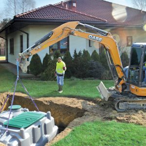 Foto des einfachen Einbaus einer Ecoflo Kleinkläranlage im Garten eines Einfamilienhauses. Es wird ein kleiner Bagger verwendet, der gerade die Anlage in die Grube hebt